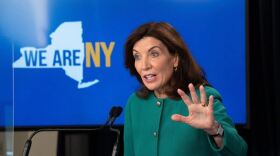 Gov. Kathy Hochul with her hand raised, wearing green, in front of a "WE ARE NY" monitor.