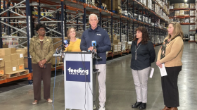 Cuatro mujeres de pie detrás de un hombre que habla en un podio, frente a un pasillo de cajas de comida.