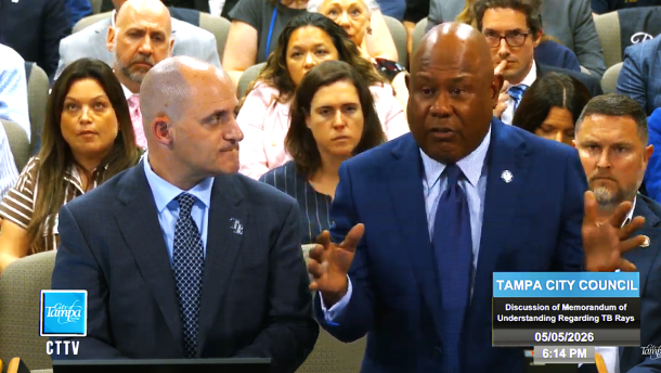 Bald white man man in a dark suit to the left looking right at a bald Black man in a blue shit speaking behind a podium with a crowd sitting behind him