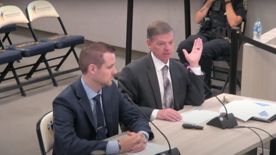 Two men sit a table in front of microphones and the one on the right speaks and gestures with his hand.