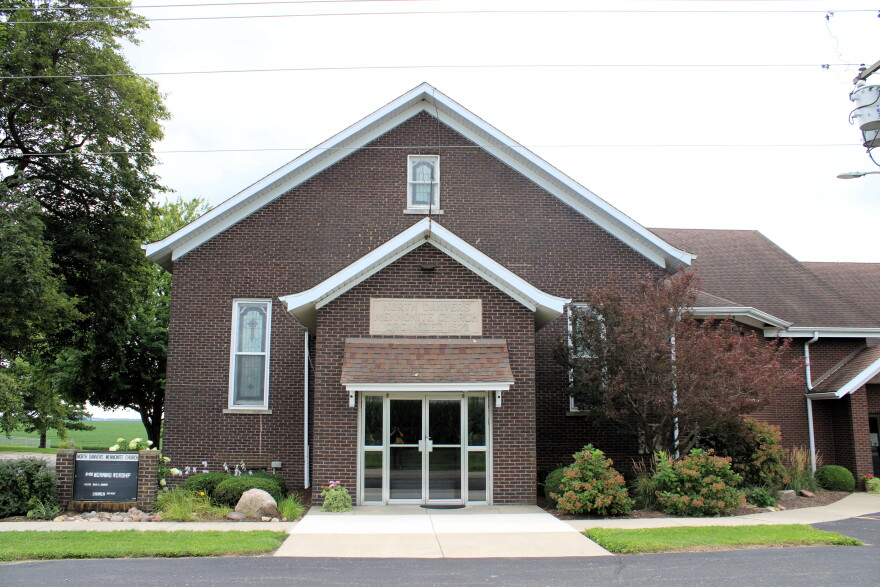 A brick church building with a glass entryway, surrounded by bushes and trees, sits near a grassy field. A black sign with white text is visible on the left side of the building.