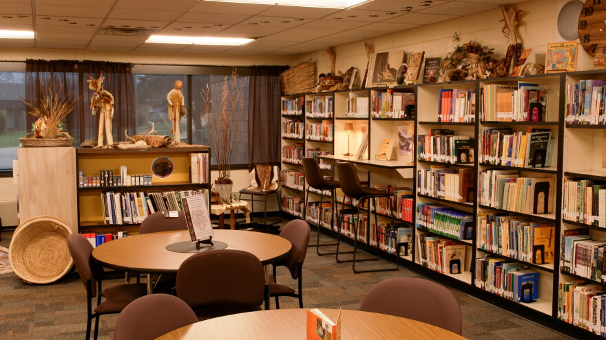 A well-lit library with round study tables, tall bookshelves filled with books, and a dedicated section featuring Indigenous and Native American cultural displays, artwork, and artifacts.