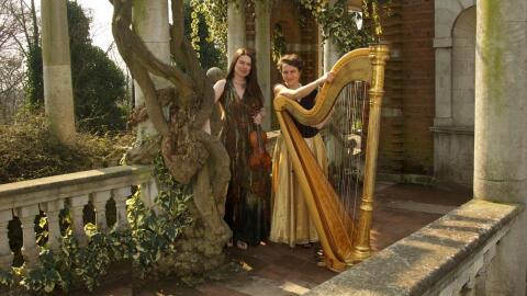 Violinist Lara St. John and harpist Marie-Pierre Langlamet standing on a sunny pathway with their instruments