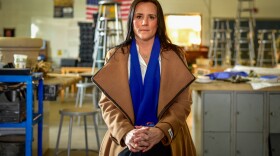 A middle-aged woman with long dark hair looks resolutely into the camera. She is seated inside a classroom.