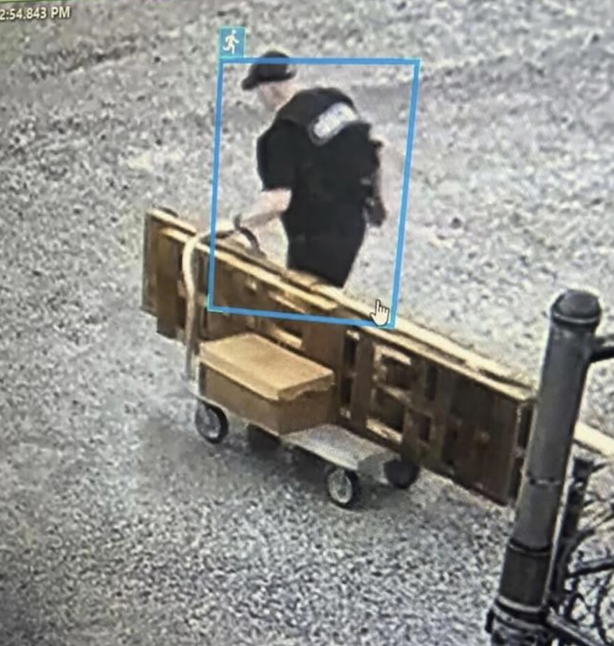 A still image taken from security camera footage shows a man, identified as Grant Hardin, seen from the rear view pushing a cart loaded with wooden pallets. The man is wearing a bulletproof vest with large white letters on the back, and has a dark hat and dark clothes on.