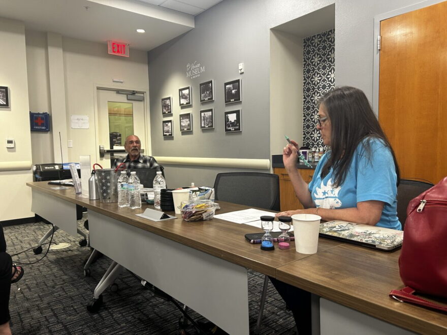 A woman with long brown hair and glasses and wearing a liight blue shirt sits at a long table during a meeting. She's holding a pen up in the air.
