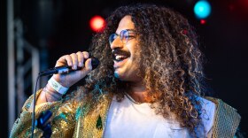 A person with long curly hair and glasses sings into a microphone
