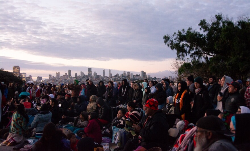 Tribal community members watch performers as the sun rises over the San Francisco skyline.
