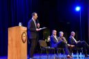 Colorado Attorney General Phil Weiser stands in a suit on stage with a microphone. Three other people sit in chairs on stage.
