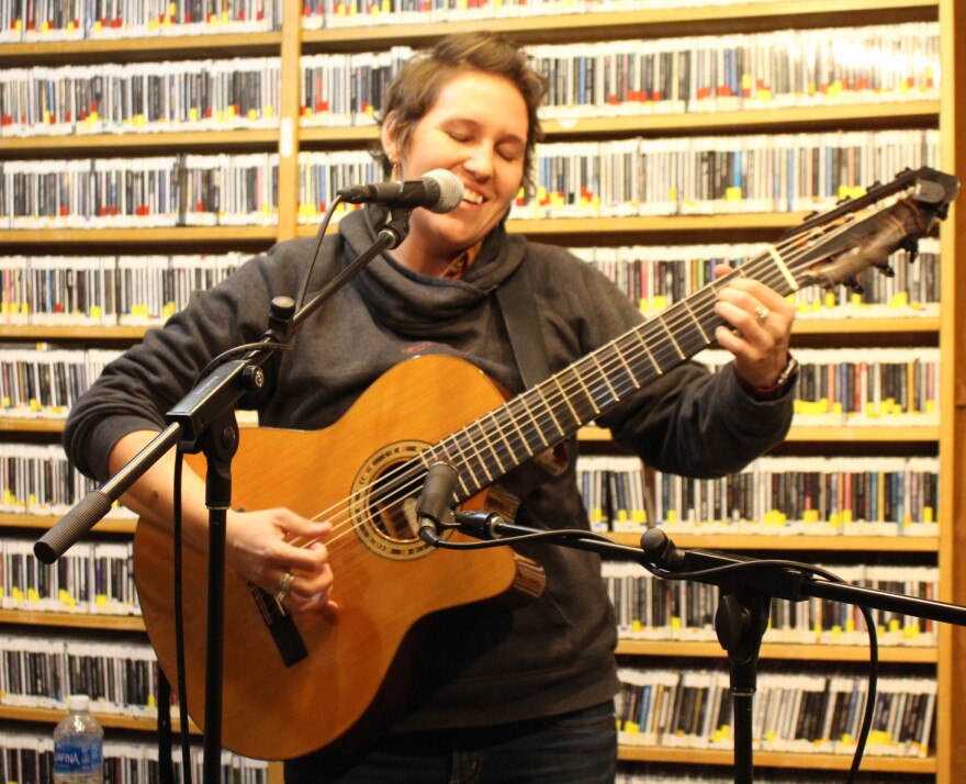 A smiling woman with her eyes closed plays guitar and sings into a microphone in a radio studio with shelves full of CDs behind her.