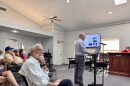 Several people look at a television with a powerpoint presentation as a man in a checkered shirt stands at a podium at the front of the room, speaking.