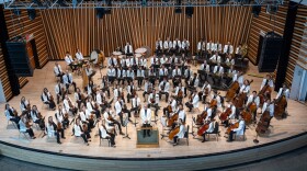 About 80 musicians who are part of the National Repertory Orchestra, are seated on a wooden stage behind the conductor. They all wear black pants and white jackets and are holding their instruments.