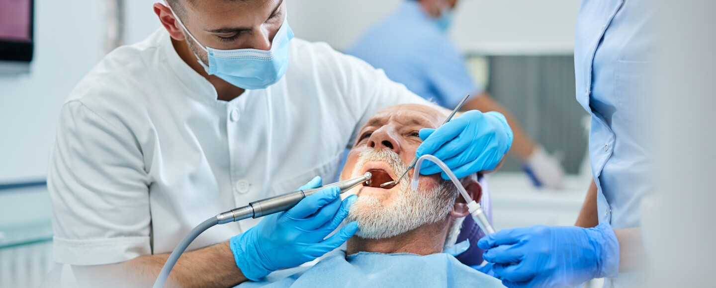 Young dentist polishing teeth of senior patient during dental procedure at clinic. Focus is on senior man.