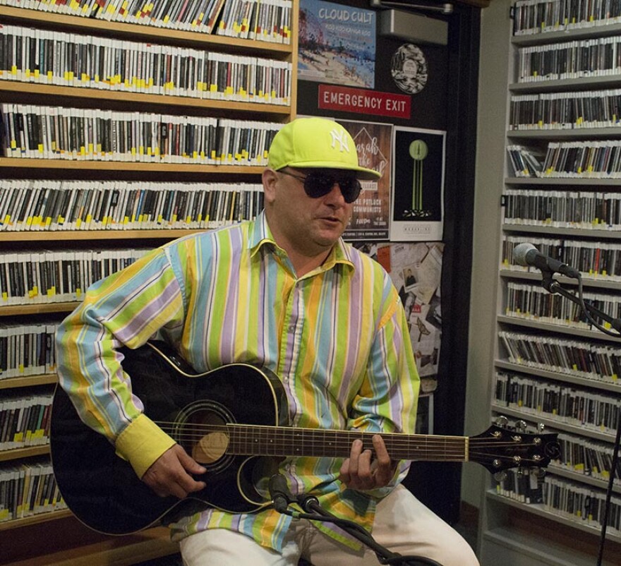 Man in bright yellow hat, brightly striped shirt, and sunglasses sitting on a stool playing guitar