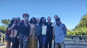 Smiling people gather around a woman in a long purple dress as she displays a proclamation.