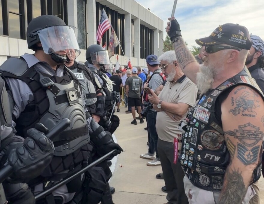Riot police in helmets and protective gear face a group of demonstrators outside a building. A tattooed man in a motorcycle-style vest raises a fist near the officers. American flags and other protesters are visible in the background.