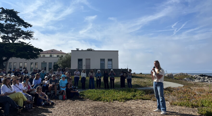 A woman stands holding a microphone with seated and standing audience members looking at her on the edge of the coast. 
