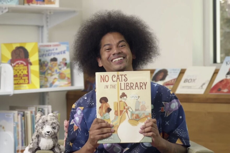 Mychal Threets, the host of "Reading Rainbow," is seen smiling and holding a book titled, "No Cats in the Library."