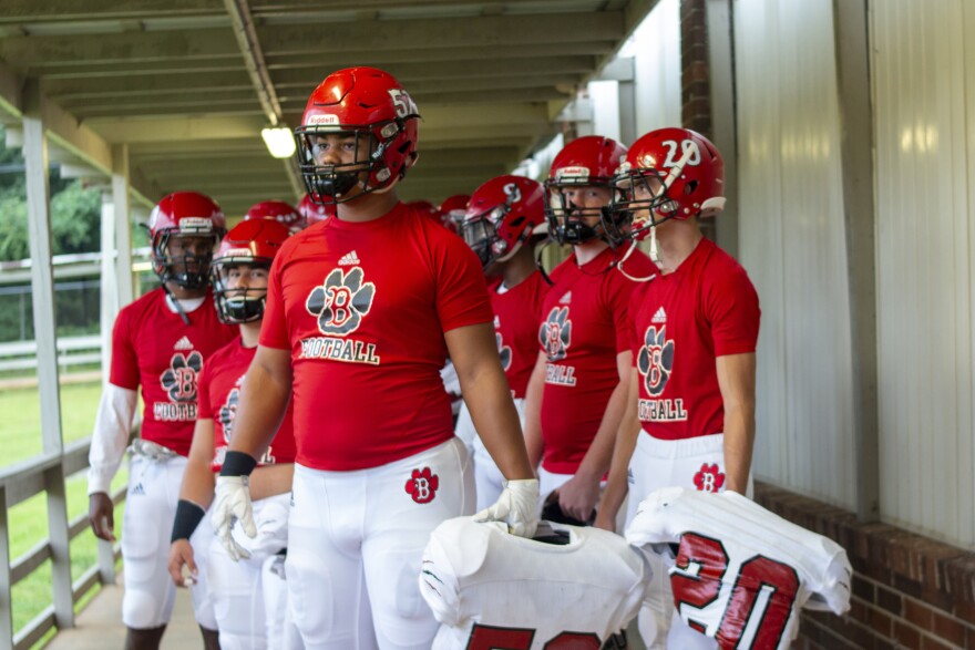 football players in red t-shirts hold their pads and look out over a field