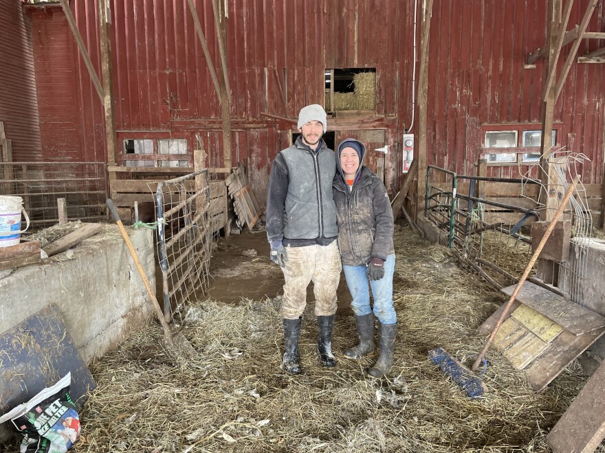 Two people stand in the entryway of a red barn with their arms around each other. They are wearing work clothes and mud boots. 