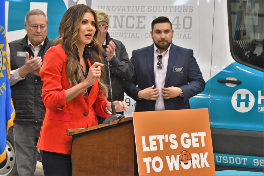 South Dakota Governor Kristi Noem speaks at a podium with a sign reading "Let's get to work" while people behind her applaud. 