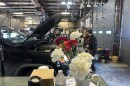 A vase of carnations in front of a car with hood open in a mechanic shop
