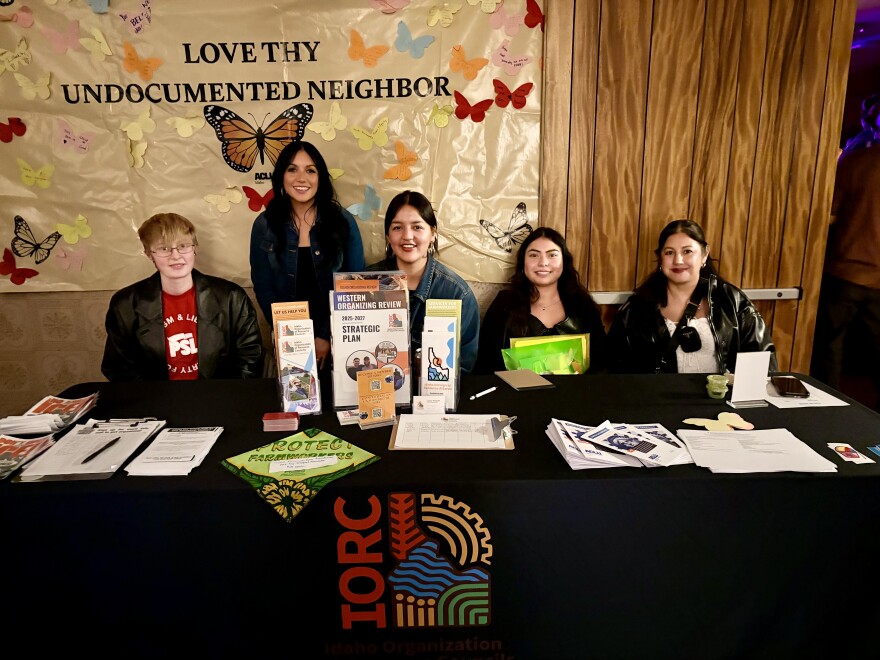Four volunteers sitting at a table with resource pamphlets for struggling families and people who want to get involved.  Jaqueline Vazquez is standing behind them and there's a poster on the wall with the words "Love thy undocumented neighbor" and butterflies of various colors.