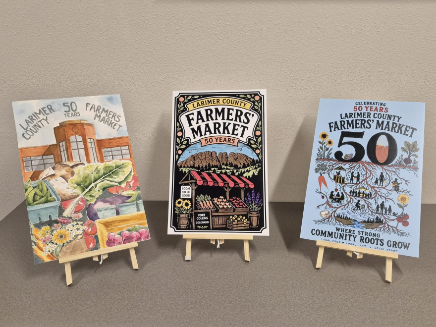 Three posters sit on tiny wooden easels. One is a watercolor painting of a brown building with vegetables in front of it. The one in the middle is a black linocut print of a market stand with vegetables for sale on it. The last one is a blue digital illustration that has roots stemming down and depictions of people and vegetables around the roots.