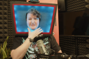 A white person with short, dirty blonde curly hair sits in a studio at a mic. They are wearing a ‘Hazbin Hotel’ t-shirt featuring Vox, a character with a TV for a head. They are also wearing a foam cosplay piece - the same TV head - that they made for a cosplay. 