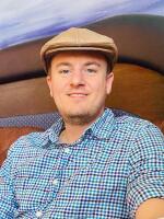 A man smiles at the camera.  He is wearing a blue checked shirt and camel colored hat.