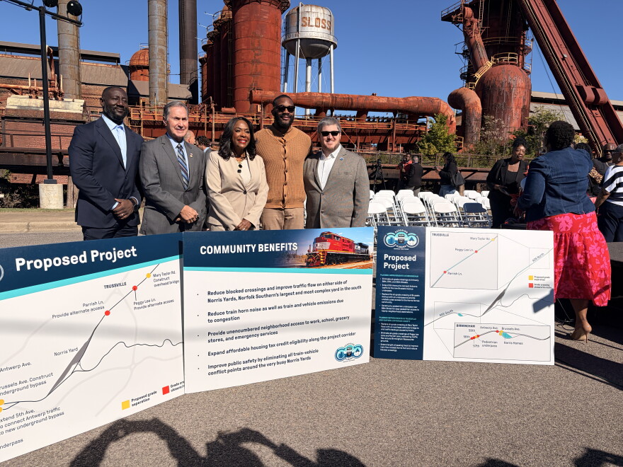 Public officials stand behind tabled-sized signs that describe the railroad project. Red industrial towers are behind them.