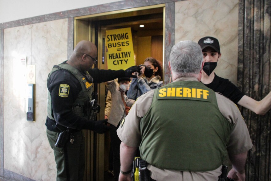 Two officers with the Jackson County Sheriff's Department usher people into an elevator. One person holds a sign that reads "strong buses = healthy city."