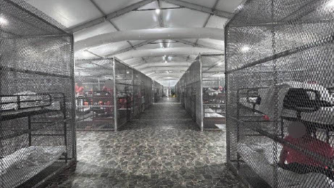 Cages filled with men in orange jumpsuits line the floor inside a tent. 