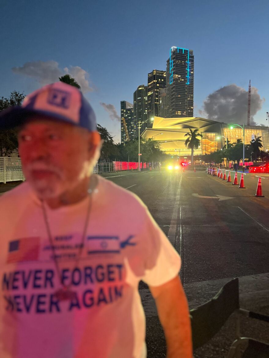 A man stands on a dark street with a lit-up venue behind him in the distance. 