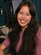 A person sits inside a radio studio. They are seated near a microphone and a computer. They are smiling and looking at the camera.