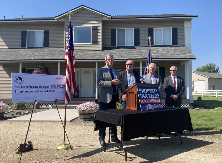 From left to right, Governor Brad Little standing in front of a house with House Speaker Mike Moyle, Representative Jason Monks and Senator Doug Ricks. An oversized check for $300 million dollars is propped on a stand to the left, next to an American flag. A poster with the words "Property Tax Relief for Idahoans" is set in front of the podium in front of the men.