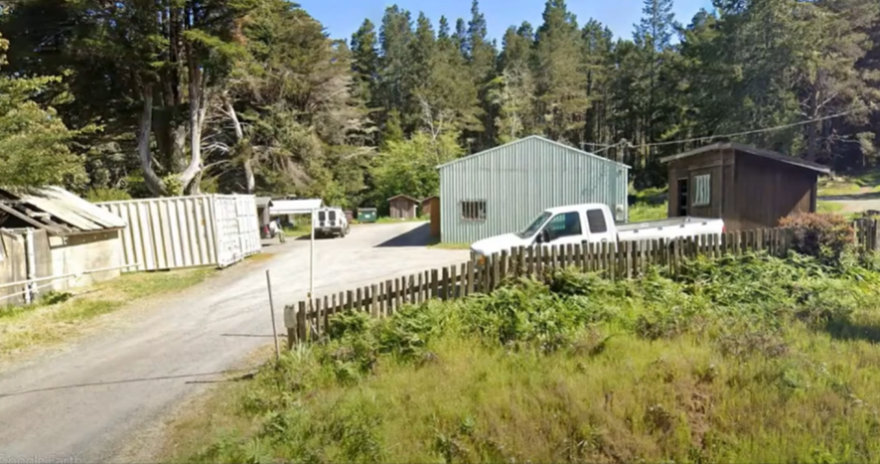 A driveway leading to a rural property with outbuildings, a shipping container, part of an unpainted picket fence, and an old white pickup truck.