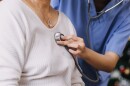 medical worker in blue scrubs holds stethoscope against chest of an elderly woman patient in white sweater top