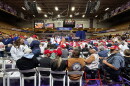 A large crowd of people seated in a high school gymnasium with a large sign in the back that reads "Make America Great Again."