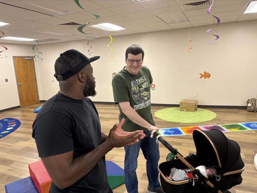 Two men talk while one holds the handle of a stroller. A baby sleeps in the stroller.