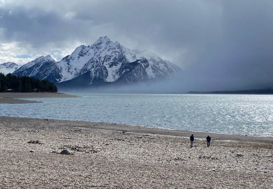 Two people stand far away the camera at Jackson Lake Dam with snowy mountains in the background.