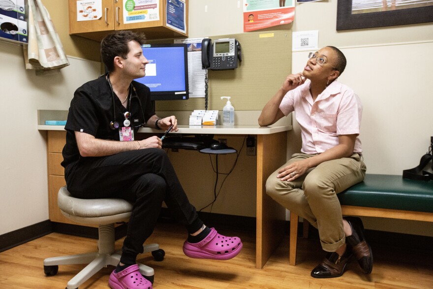 a physician wearing black scrubs and bright pink Crocs speaks to a patient sitting on a bench in an exam room