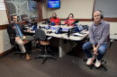 Four smiling people wearing headphones sit at a table in a radio talk studio: a man front left has short brown hair and a brown mustache and beard and is wearing glasses, a dark blue button-down shirt and lighter blue tie under a black suit coat, khakis, dark socks, and brown shoes; a man back left has short black hair and a black mustache and beard and is wearing a red polo shirt; a woman back center has short brown hair and is wearing glasses and a red graphic t-shirt; a man at right has very short dark hair and is wearing glasses, a purple and white checked button-down shirt, jeans, and white and red sneakers