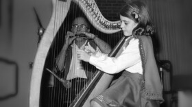 A young girl plays an Irish harp as her father plays harmonica in the background