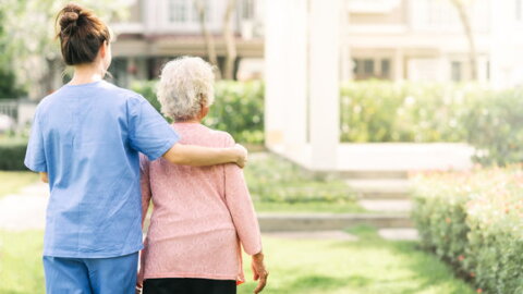 woman in blue scrubs has her arm an older woman with white hair as she escorts her over a grassy green courtyard area, both faces turned away from camera