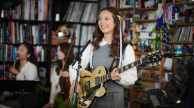 A young woman smiles while standing behind an office desk and playing guitar with a microphone in front of her and two woman playing stringed instruments in the background.