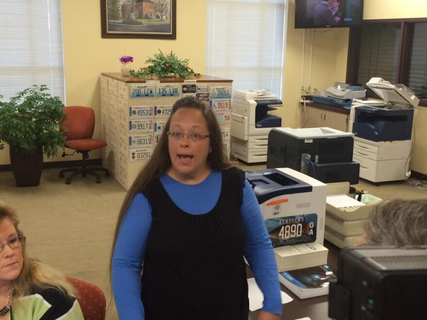 A woman in a blue shirt and black jumper looks toward the camera with her mouth open as if to speak. She's in an office setting. 