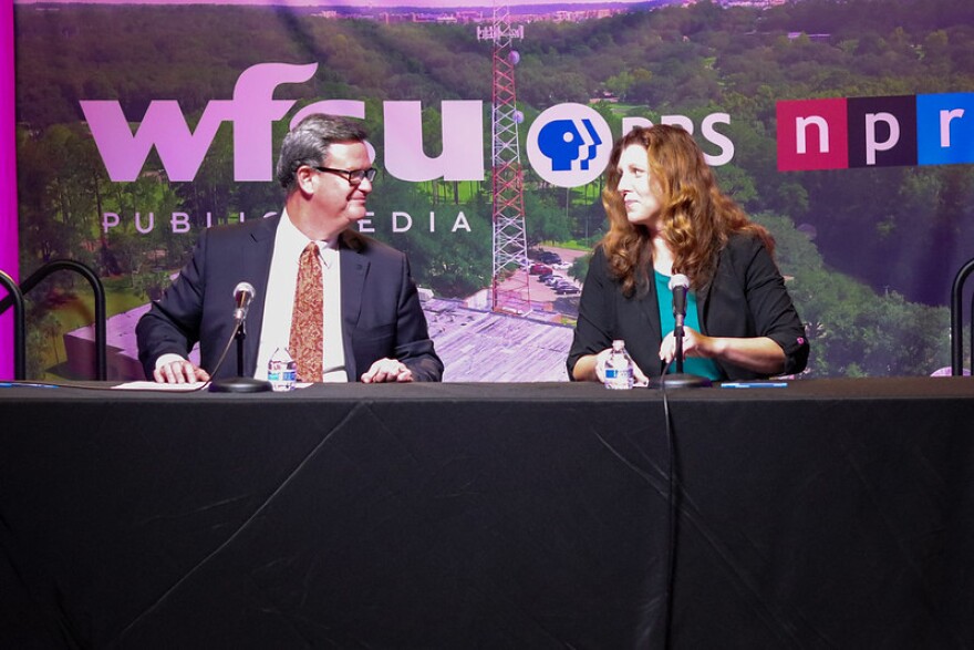 A man in a suit and tie and a woman with long hair sit behind microphones at a table, looking at each other. The banner behind them says "WFSU" and "NPR" and has a photo of a radio tower. 