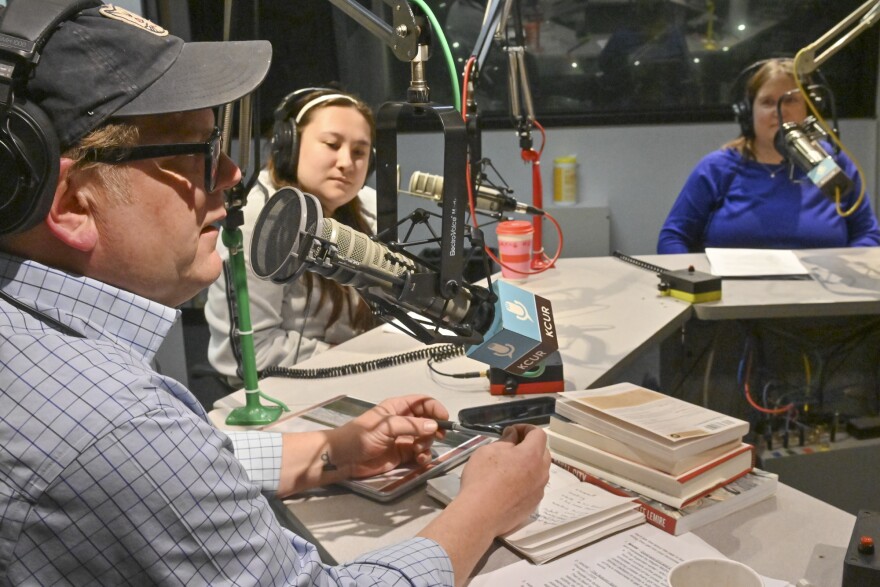 Three people are seated inside a radio studio. They are sitting behind microphones. The man at left, is talking and holding a pen in both hands with a small stack of books in front of him.