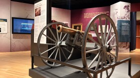 A museum exhibit with informative (but not readable at distance) panels lining the walls. At the center of the exhibit is a brass cannon mounted on a wooden unit with a hitch and two large wheels.
